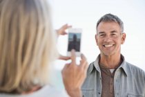 Woman taking photo of husband with cell phone on beach — Stock Photo