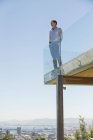 Hombre de pie en la terraza con valla de vidrio con las manos en los bolsillos - foto de stock