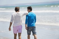 Расслабленные мужчины, идущие по пляжу с волнистым морем — стоковое фото