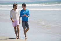 Homens sorridentes andando na praia de areia com mar ondulado — Fotografia de Stock