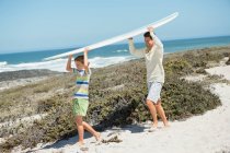 Hombre y su hijo llevando una tabla de surf en la playa - foto de stock