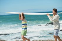 Man and his son carrying a surfboard on the beach — Stock Photo