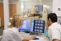 Médicos examinando a varredura no computador com o paciente no scanner de ressonância magnética em segundo plano — Fotografia de Stock
