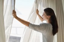 Lächelnde Frau öffnet Vorhang des Fensters zu Hause — Stockfoto