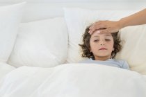 Human hand examining fever of little boy lying in bed — Stock Photo