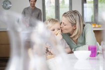 Mujer desayunando con su hijo pequeño en la cocina con su marido en el fondo - foto de stock