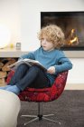 Garçon lisant un livre dans un fauteuil dans le salon à la maison — Photo de stock