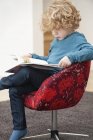 Милий хлопчик з світлим волоссям читає книгу в кріслі вдома — стокове фото