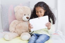Happy little girl using digital tablet with teddy bear on bed — Stock Photo