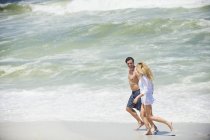 Happy couple running on beach with wavy sea on background — Stock Photo