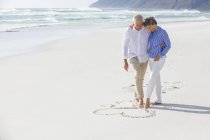Abrazando pareja mayor dibujo forma de corazón en la arena con los pies - foto de stock