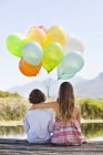 Rear view of siblings sitting with colorful balloons at wooden pier at lake in nature — Stock Photo