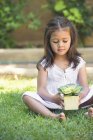 Симпатична маленька дівчинка тримає рослину в горщику, сидячи на траві — стокове фото