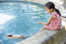 Маленькая девочка сидит на краю бассейна и играет с игрушечной лодкой в воде — стоковое фото
