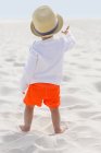 Vista trasera del niño en sombrero de paja de pie en la playa de arena - foto de stock