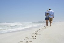 Vista traseira do casal romântico andando na praia sob o céu azul com pegadas na areia — Fotografia de Stock