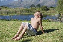 Niño sin camisa sentado con los ojos cerrados en la hierba contra las montañas - foto de stock