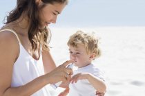 Mulher pulverizando protetor solar na mão do bebê na praia — Fotografia de Stock