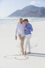 Embracing senior couple drawing heart shape on sand with feet — Stock Photo