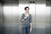 Donna d'affari in possesso di tazza usa e getta e sorridente davanti all'ascensore — Foto stock