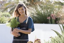 Portrait of young woman in apron holding flower pot in garden — Stock Photo