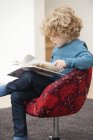 Lindo chico con pelo rubio leyendo un libro en sillón en casa - foto de stock