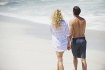 Rückansicht eines Paares, das Händchen haltend am Strand spaziert — Stockfoto