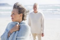 Nahaufnahme einer nachdenklichen Seniorin, die am Strand steht, mit Ehemann im Hintergrund — Stockfoto