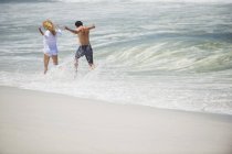 Visão traseira de casal alegre correndo em onda na praia — Fotografia de Stock