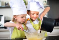 Двое маленьких детей счастливая детская семья мальчика и девочки с фартуком и шляпой шеф-повара, которые готовят веселые кулинарные блюда на кухне дома. — стоковое фото