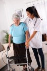 Весела молода медсестра реабілітації, яка допомагає літній старшій жінці, використовуючи ходунки — стокове фото