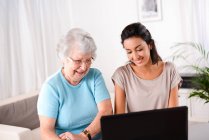 Joyeuse jeune femme aidant une personne âgée utilisant un ordinateur portable pour la recherche sur Internet et e-mail — Photo de stock