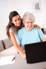 Joyeuse jeune femme aidant une personne âgée utilisant un ordinateur portable pour la recherche sur Internet et e-mail — Photo de stock