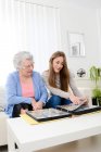 Mulher idosa com sua jovem neta em casa olhando para a memória no álbum de fotos da família — Fotografia de Stock