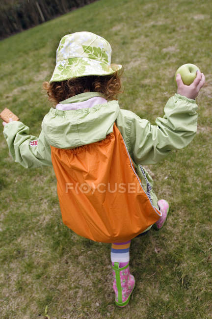 Little girl in hat and rain-jacket with backpack walking in garden and holding apple and cake — Stock Photo
