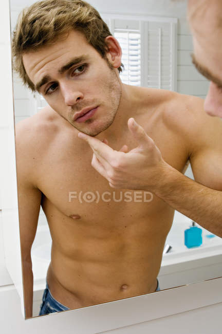 Hemdloser junger Mann prüft Gesicht vor Badezimmerspiegel — Stockfoto