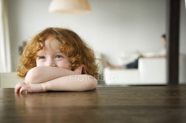 Веселая рыжая маленькая девочка сидит за журнальным столиком и смотрит в сторону — стоковое фото