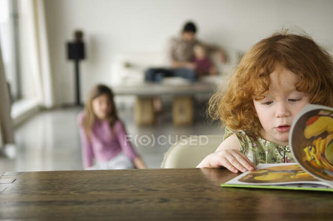 Bambina che legge il libro per bambini, la famiglia sullo sfondo — Foto stock