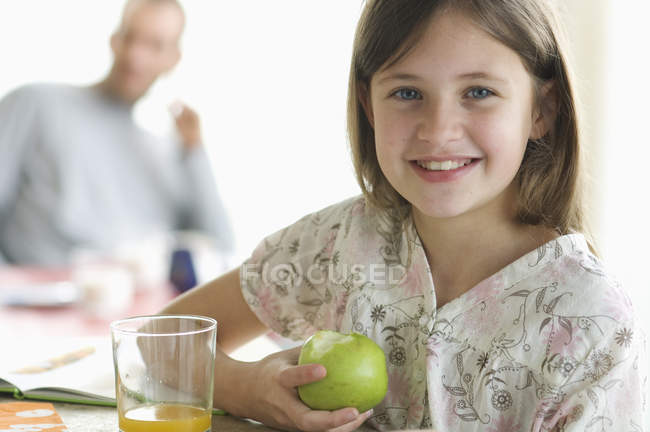 Retrato de niña sonriente comiendo manzana en la mesa con el hombre en el fondo - foto de stock