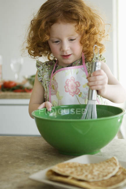 Pequeña chica de jengibre mezclando ingredientes en un tazón con un batidor en la cocina - foto de stock