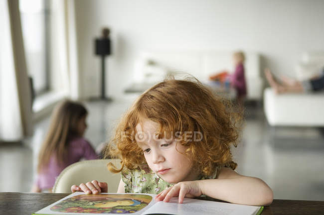 Pequeña chica pelirroja leyendo libro en la mesa en casa - foto de stock