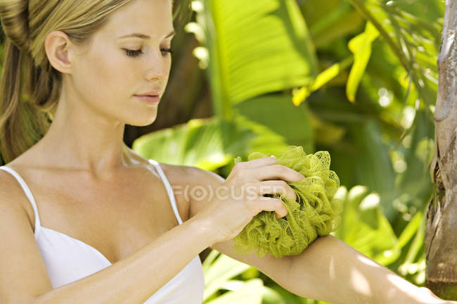 Portrait de jeune femme nettoyage bras avec maille éponge à l'extérieur — Photo de stock