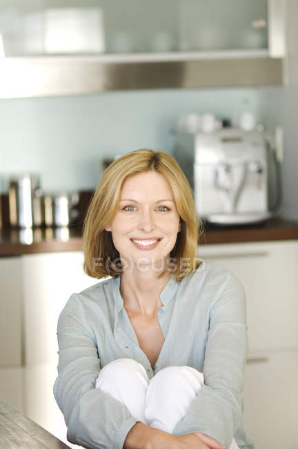 Улыбающаяся вдумчивая женщина сидит на кухне и смотрит в камеру — стоковое фото