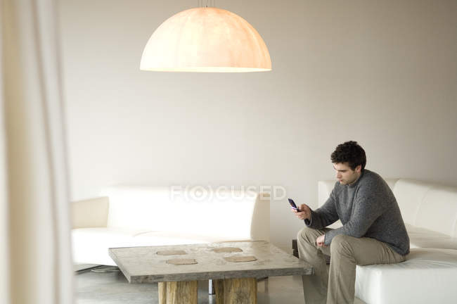 Мужчина сидит на диване в гостиной и пользуется мобильным телефоном — стоковое фото