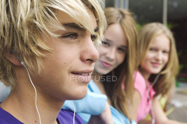 Portrait of teenage boy with earphones, 2 teenage girls in background — Stock Photo