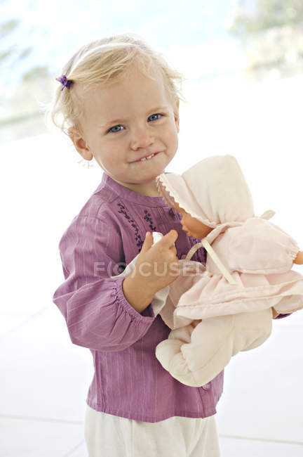 Маленькая девочка смотрит в камеру и держит куклу на размытом фоне — стоковое фото