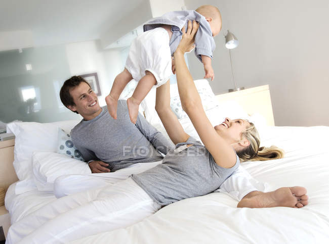 Веселые родители и ребенок веселятся на кровати в спальне, женщина поднимает ребенка — стоковое фото
