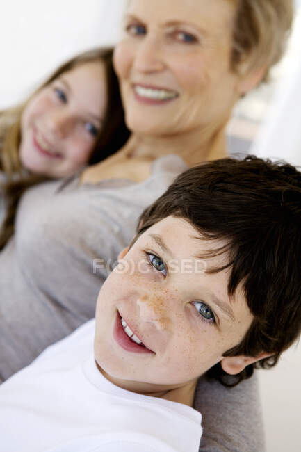 Mujer mayor y dos niños sonriendo para la cámara, en el interior - foto de stock