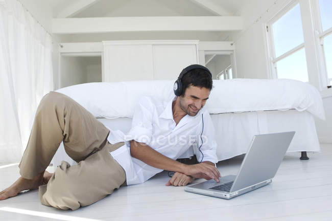 Молодой улыбающийся мужчина использует ноутбук, лежа рядом с кроватью — стоковое фото