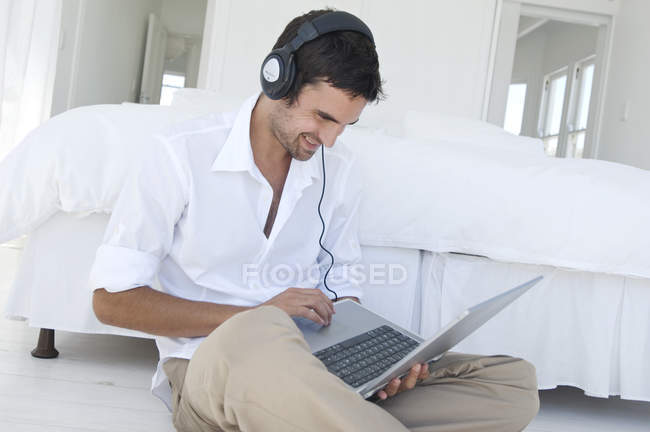 Молодий усміхнений чоловік використовує ноутбук, лежачи біля ліжка — стокове фото
